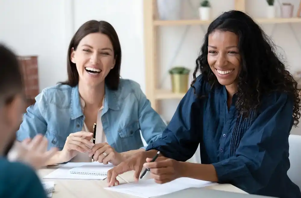 Two women share a joyful moment, laughing together during a meeting, highlighting camaraderie and positive engagement.